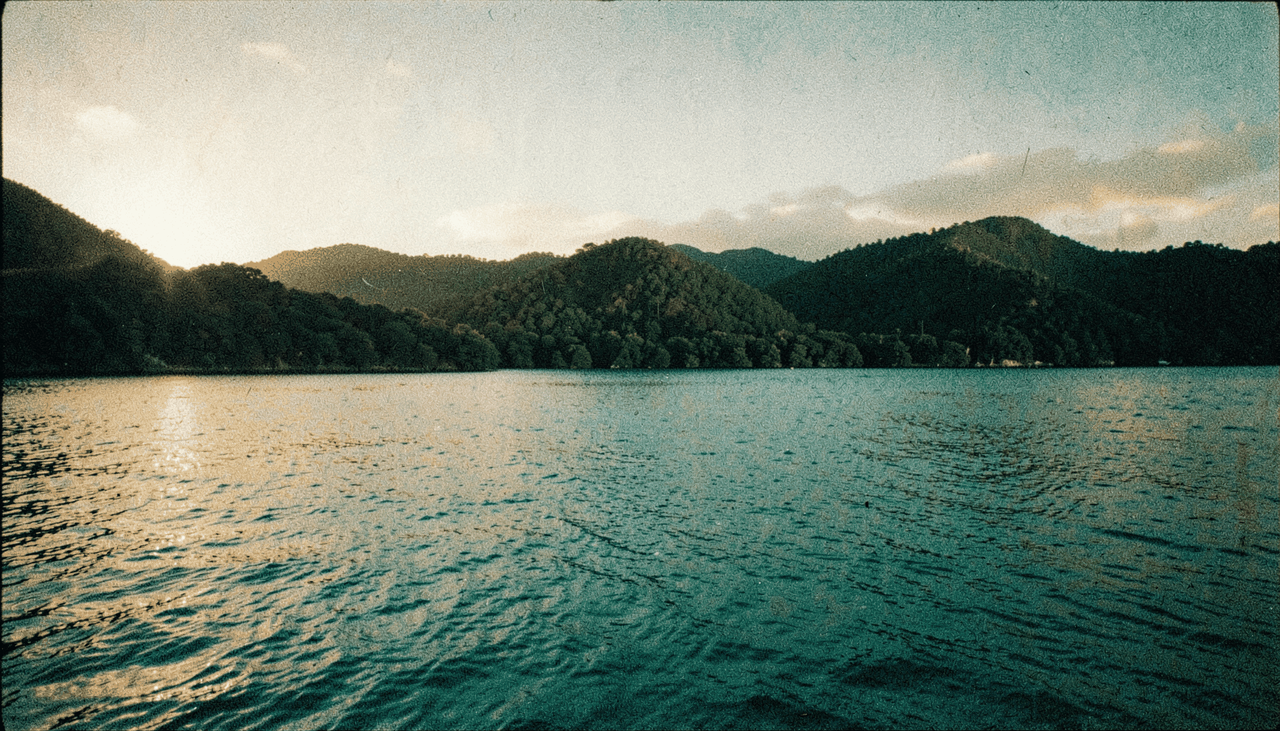 Photographie argentique granuleuse d'un lagon polynésien au coucher du soleil, tons turquoise profond et or doux, collines verdoyantes en arrière-plan